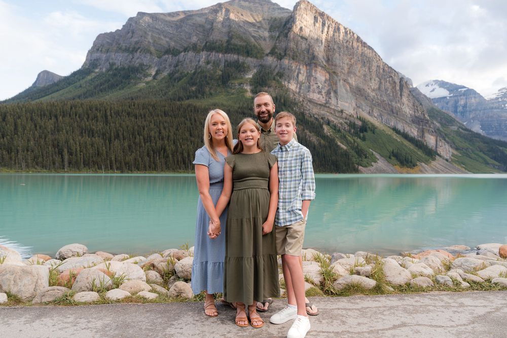 Family of four posing at turquoise Lake Louise with Rocky Mountain peaks and evergreen forest backdrop.