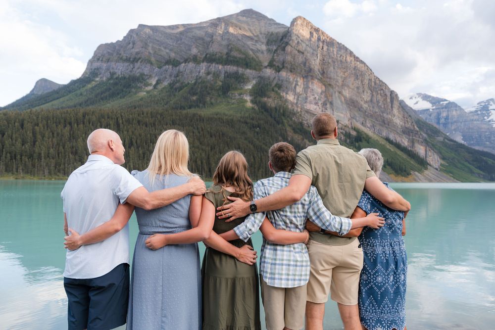Family group standing arm in arm facing turquoise mountain lake with rocky peaks and evergreen forests in background.