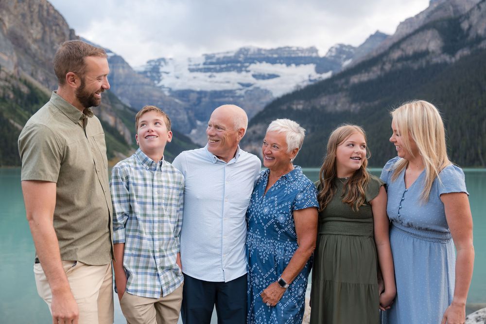 Family of six smiling together at Lake Louise with turquoise water and snow-capped mountains in background.