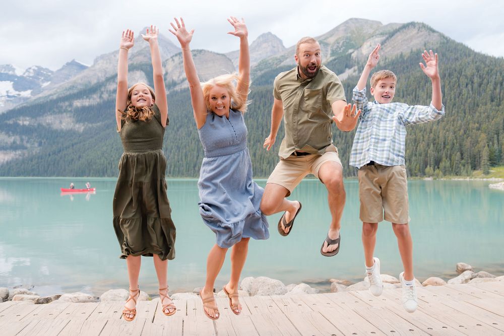 Family of four jumping happily on a dock with a stunning turquoise alpine lake and mountains behind them.