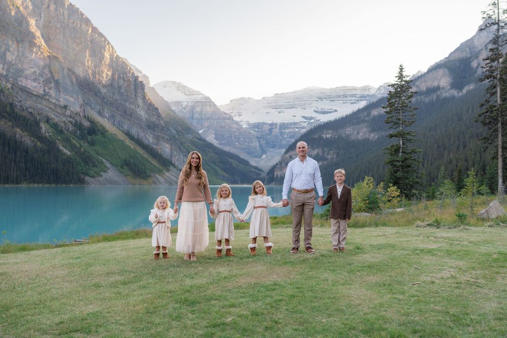 Family of six holding hands at Lake Louise, with turquoise water and snow-capped mountains in background.