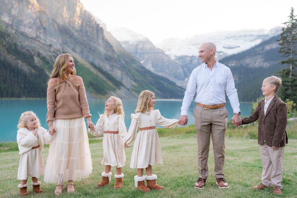 Family of six holding hands in front of turquoise Lake Louise with mountain backdrop on a sunny day.