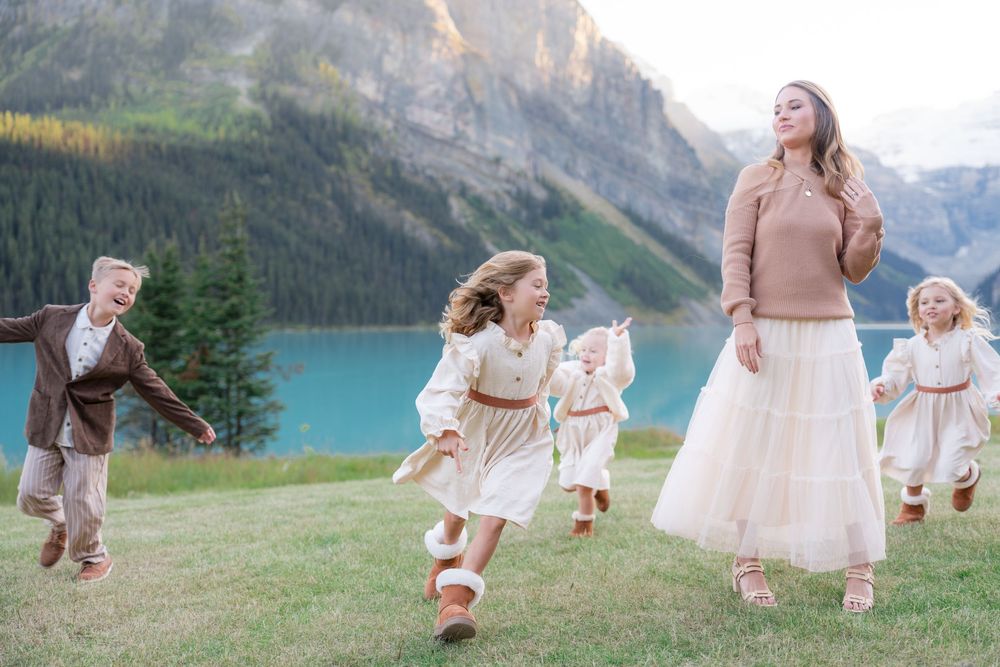 A woman and children run joyfully on a green lawn with a stunning turquoise lake and mountain backdrop.