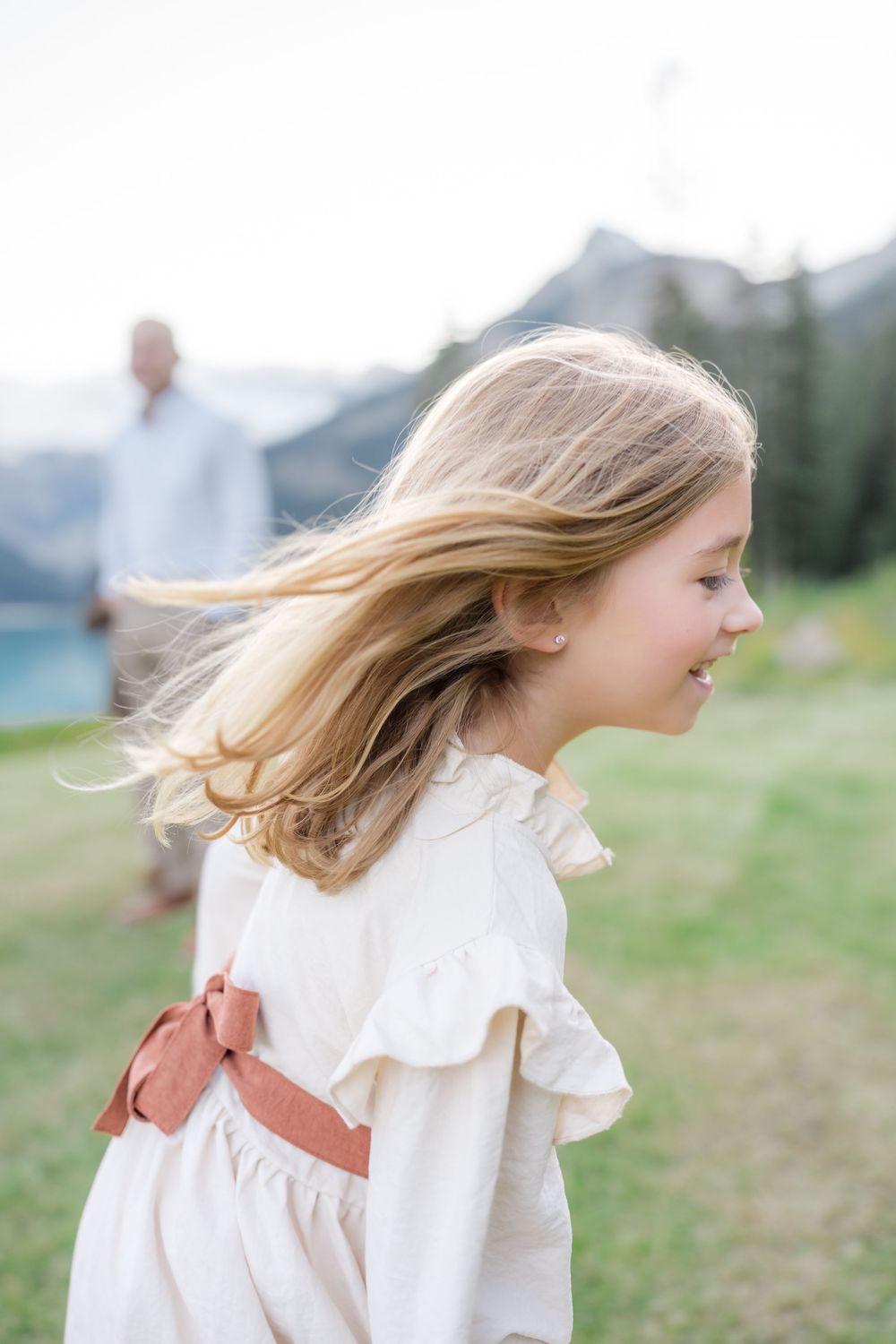 Young girl in white ruffled dress with pink bow, hair blowing in wind, smiling outdoors near mountain lake.