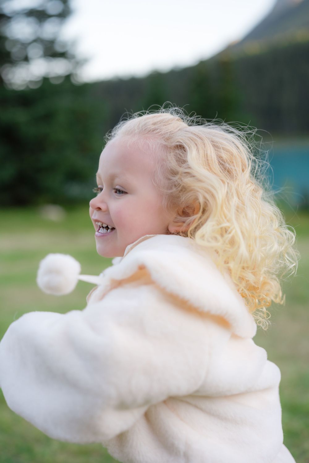 Curly blonde toddler girl laughing outdoors in white fluffy coat near mountain lake.