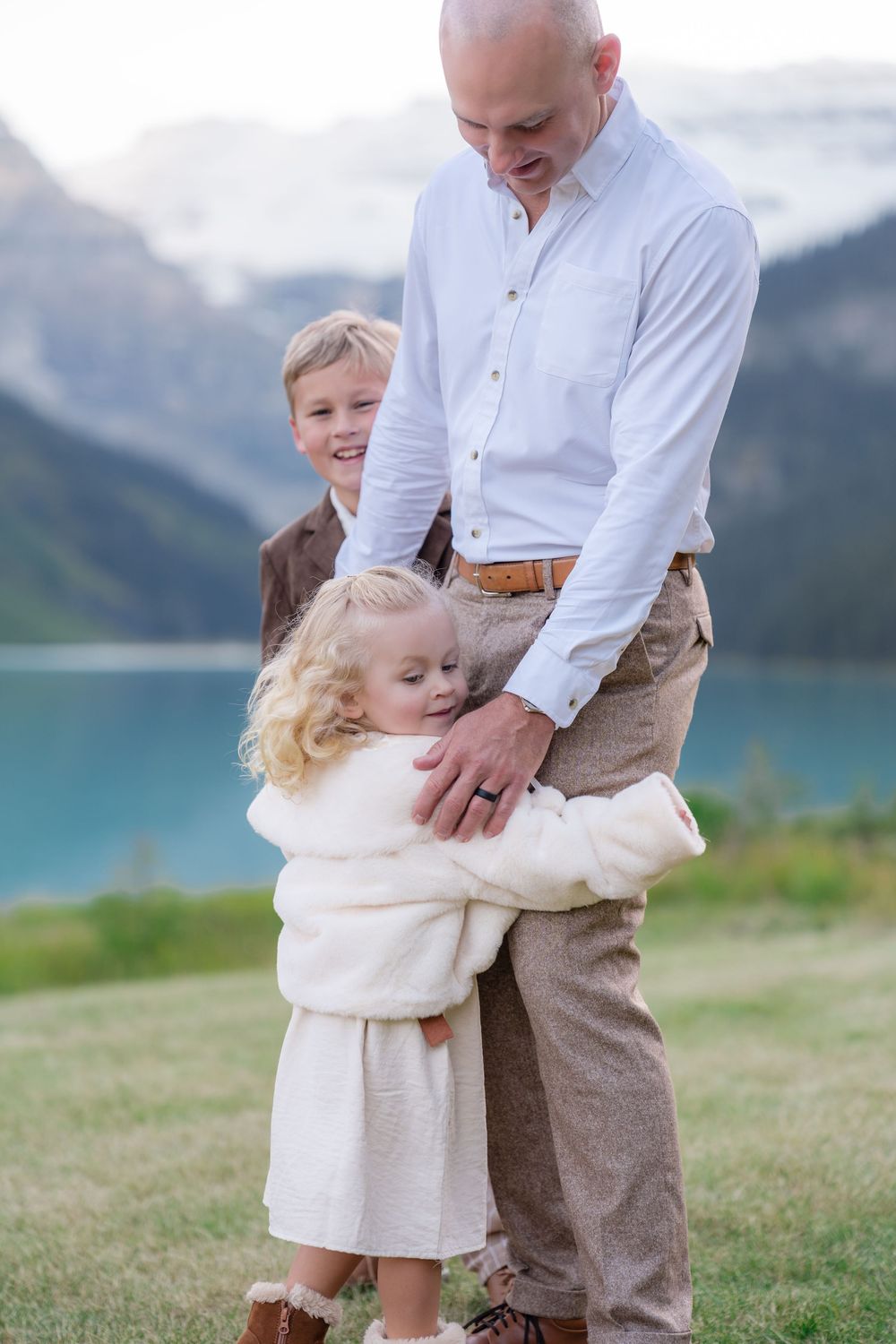 Bald father smiles as young daughter hugs him and son stands beside him at Lake Louise.