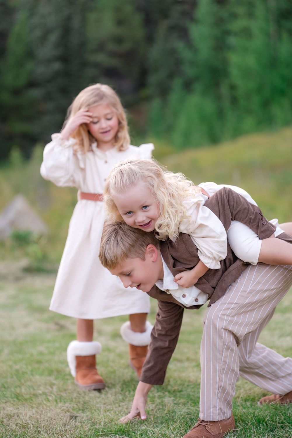 Boy giving piggyback ride to curly-haired girl while another blonde girl watches outdoors on green grass.