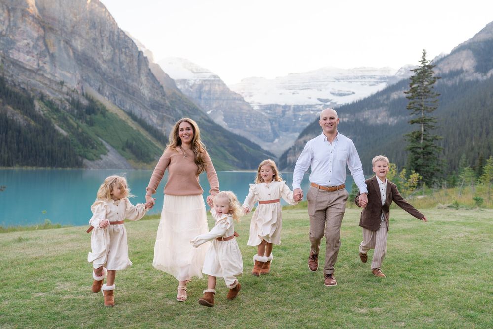 Family of six walking hand-in-hand by turquoise Lake Louise with snow-capped mountains in background.