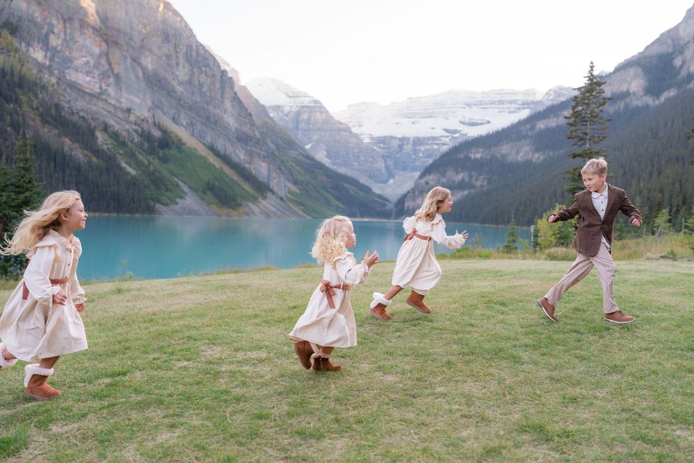 Four children run and play on green grass with turquoise Lake Louise and mountains behind.