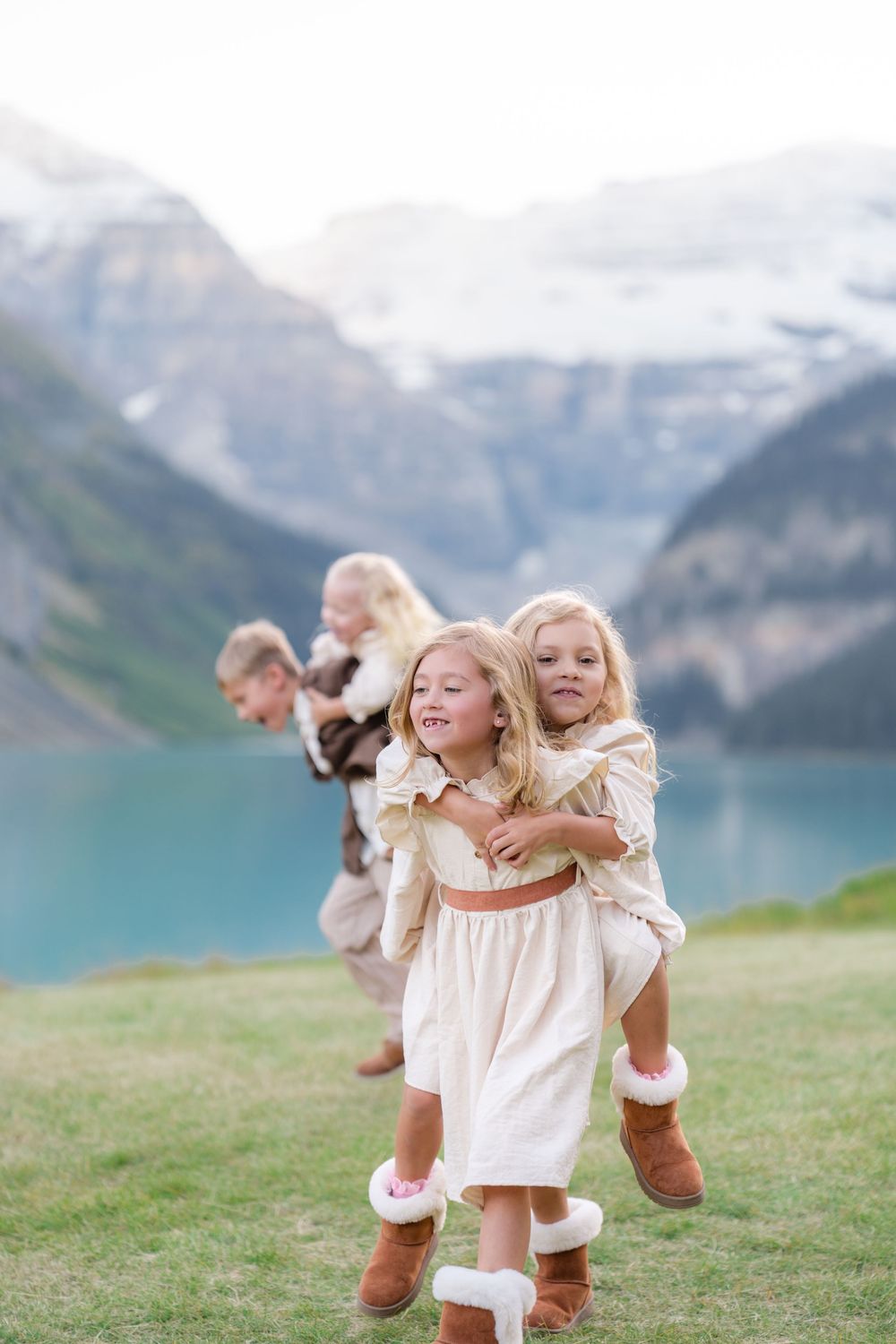 Girls in cream dresses and UGG boots play piggyback near turquoise Lake Louise with snow-capped mountains backdrop.
