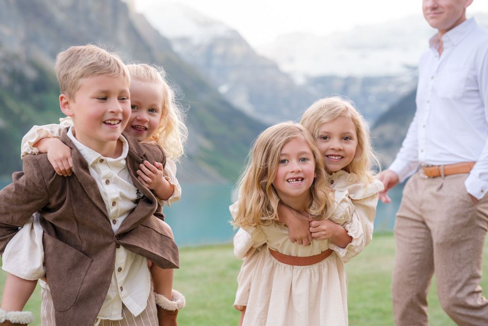 Children laughing and playing piggyback near a scenic mountain lake, dressed in elegant cream and brown outfits.