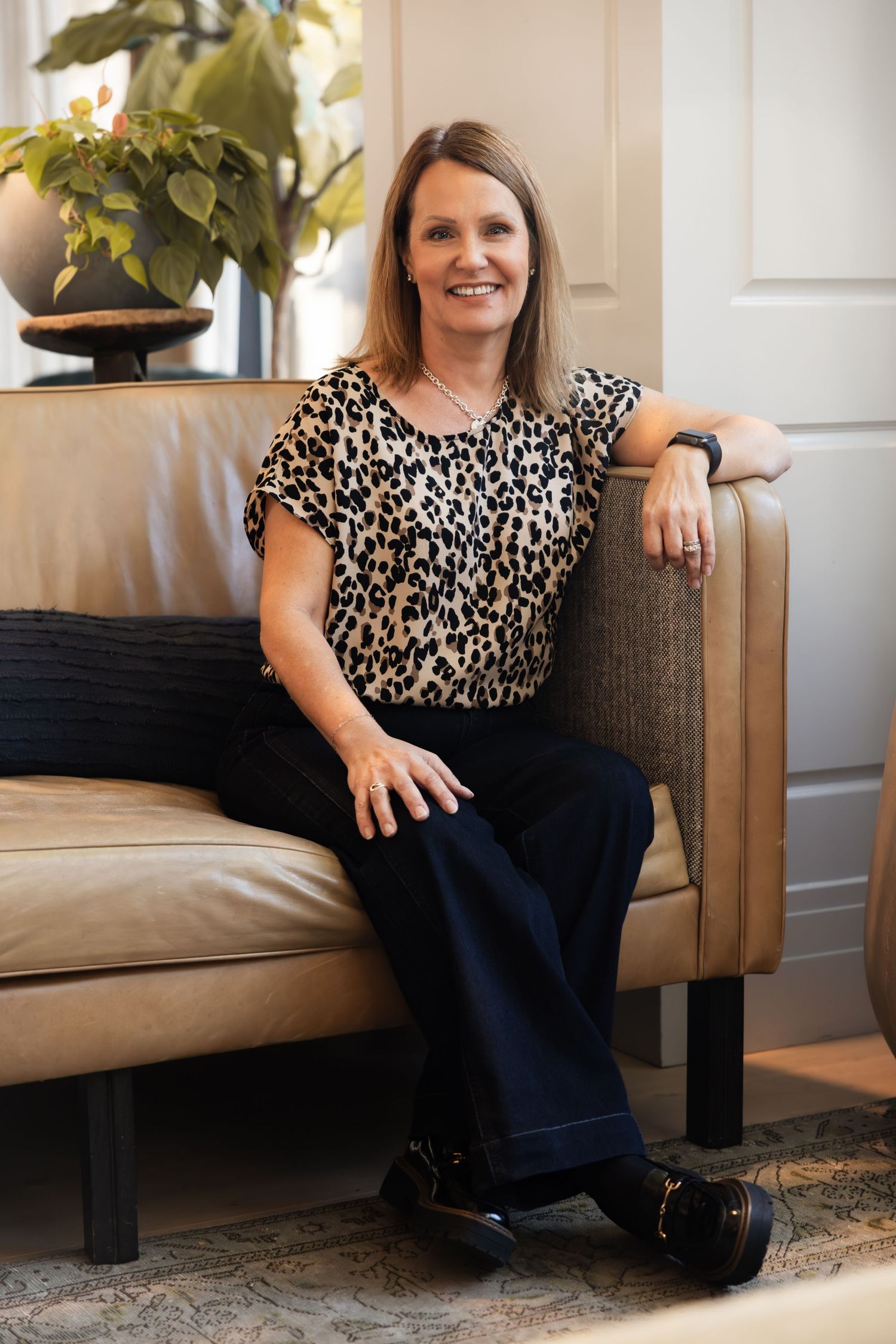 A person in a leopard print top and dark pants sits comfortably on a tan couch with a houseplant visible in the background.