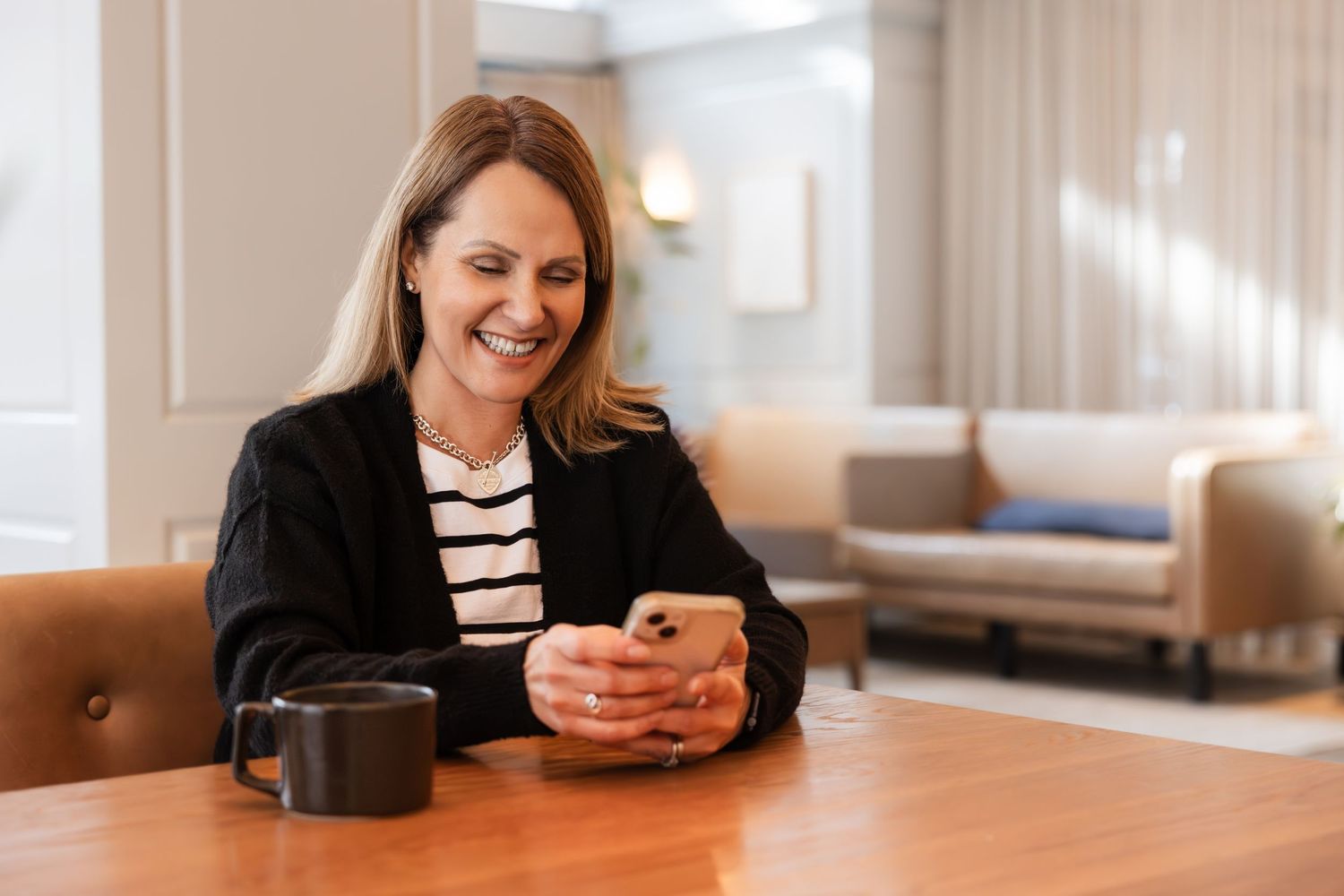 Professional looking at smartphone while sitting at wooden table with black coffee mug in bright modern office space.