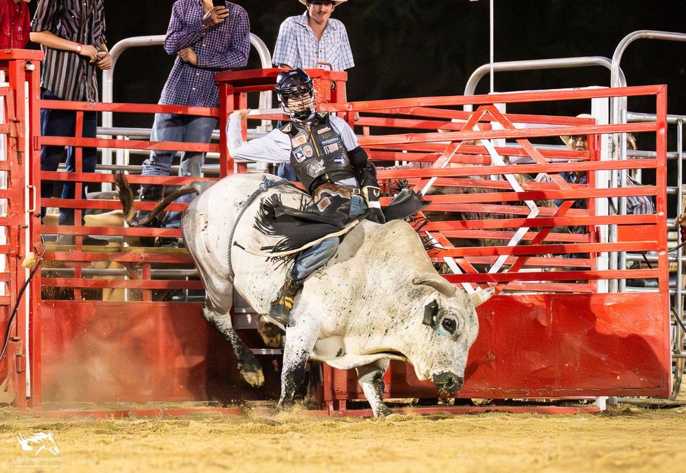 A bull rider gets thrown from a bucking bull during a rodeo competition in an arena with red gates.