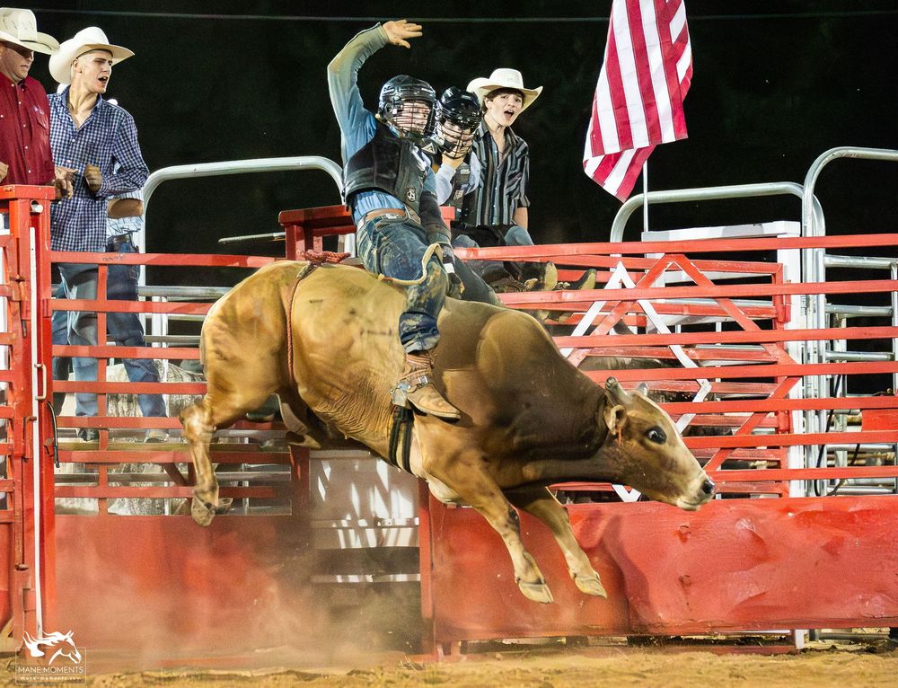 A bull rider competes in a rodeo arena with a bucking bull while an American flag waves in the background.