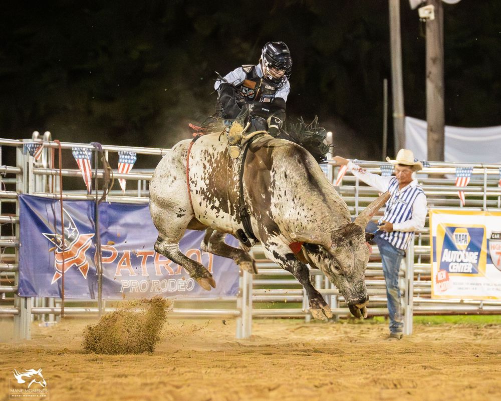A bull rider competes in a nighttime rodeo event as the bucking bull kicks up dirt in an illuminated arena.