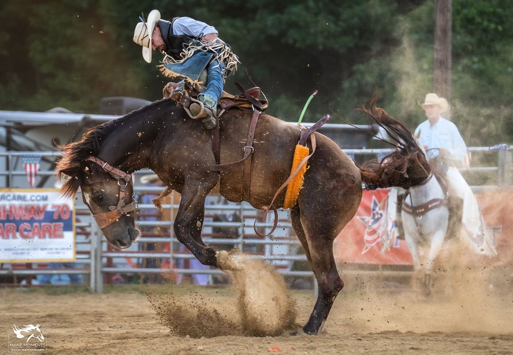 A bronc rider competes in a rodeo arena as the horse bucks and kicks up dust while officials look on from behind.