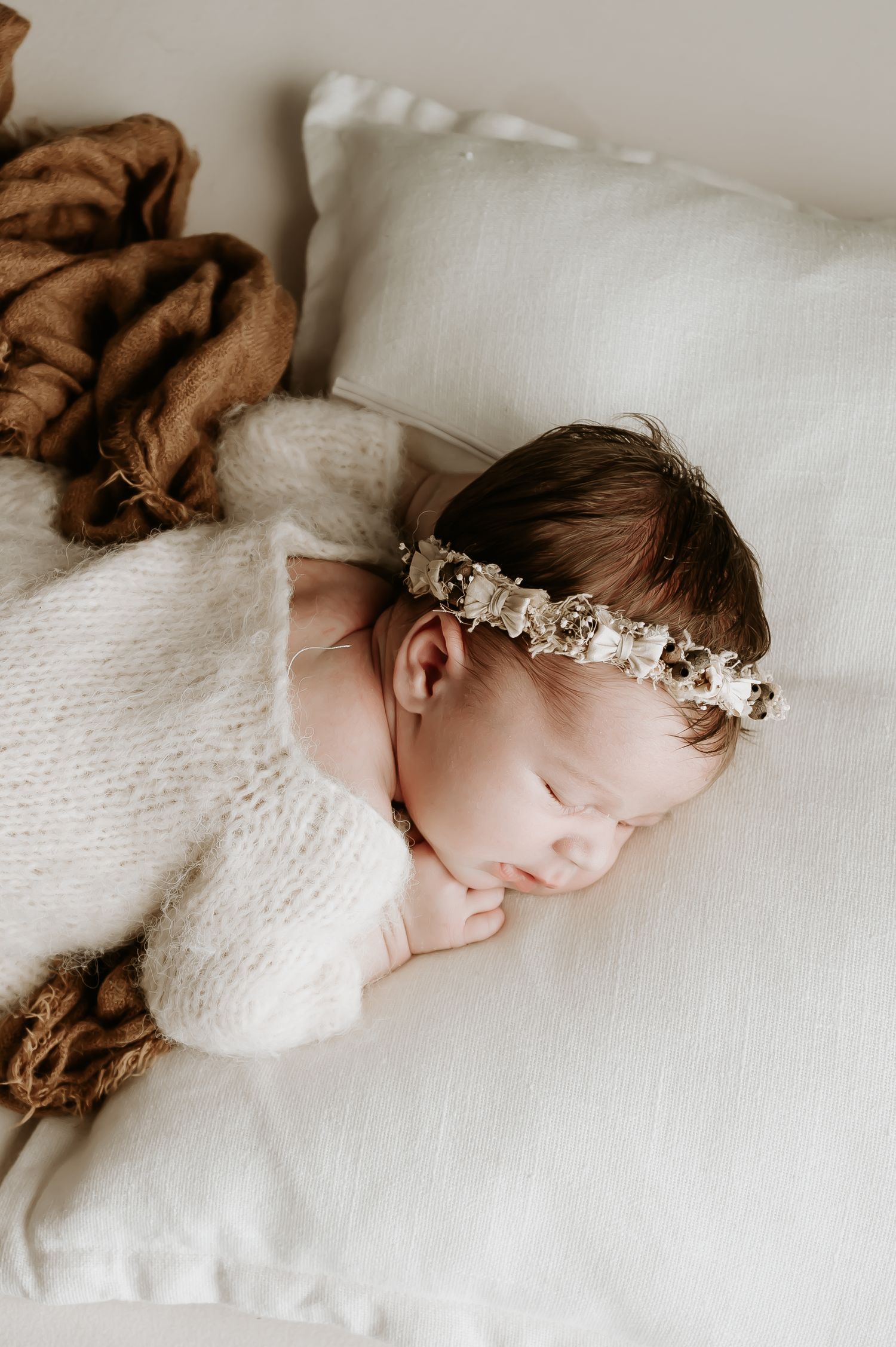 Sleeping newborn baby wearing delicate floral headband on white bedding.