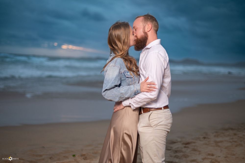 Couple sharing a romantic kiss on the beach at sunset, with waves in the background and a moody blue sky overhead