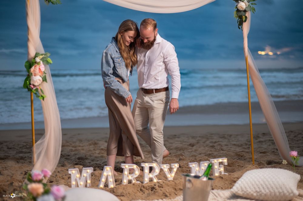 Romantic beach proposal setup at sunset with a couple standing under a canopy, surrounded by flowers and decor, with ‘MARRY ME’
