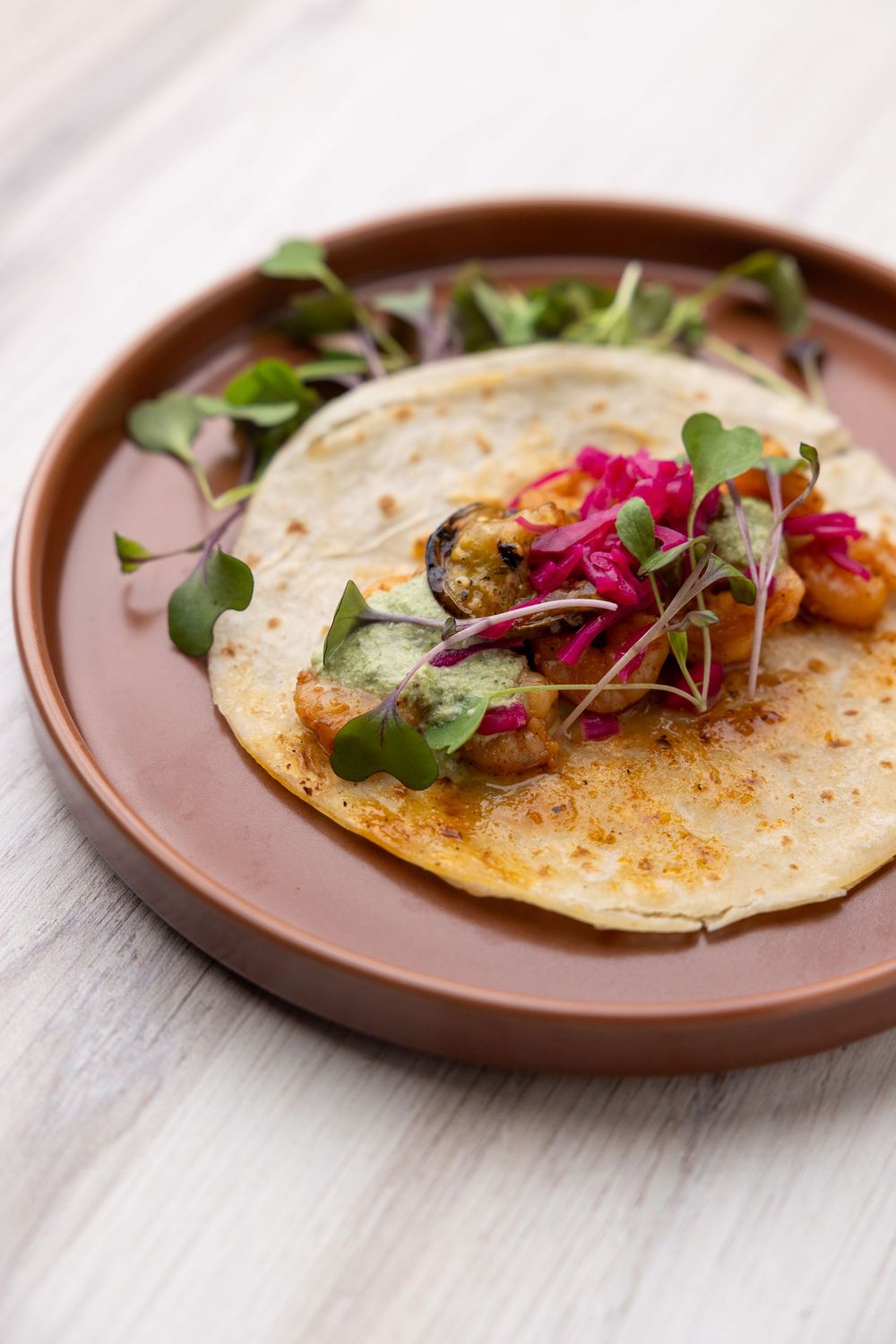 Gourmet taco topped with fresh microgreens and pink edible flowers on a rustic brown plate.