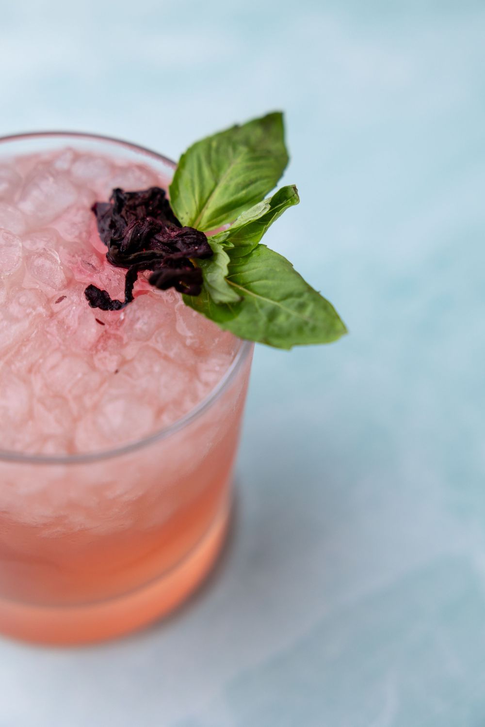 Pink cocktail with crushed ice, fresh basil leaf and dark berry garnish in a lowball glass.