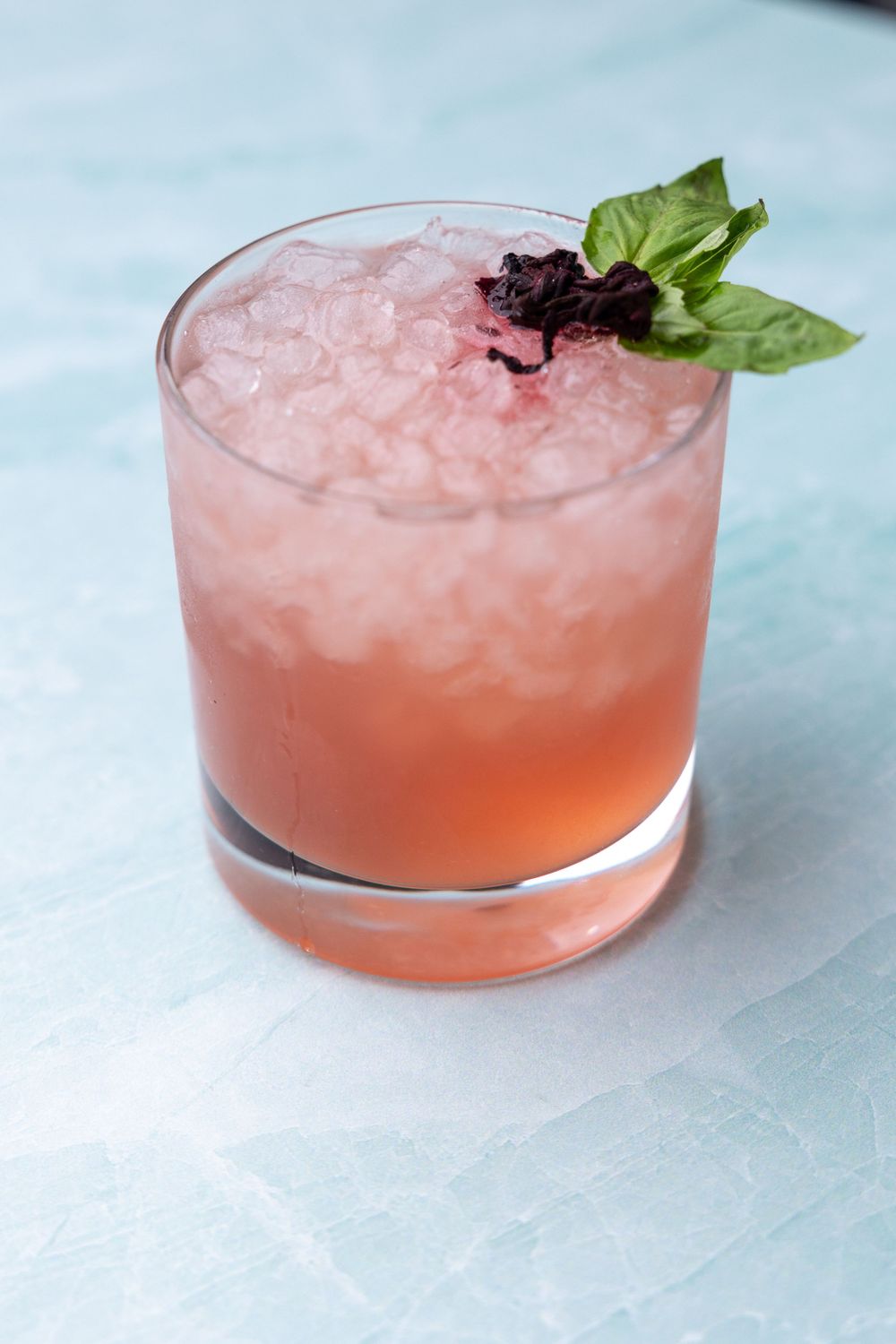 Refreshing pink cocktail with crushed ice and fresh herb garnish on a light blue surface.