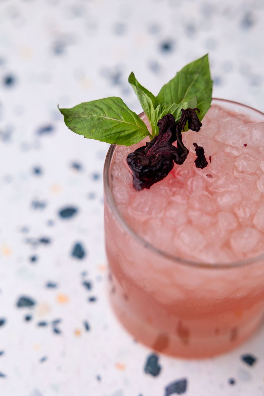 Pink cocktail garnished with fresh basil and dark berries on a speckled surface.