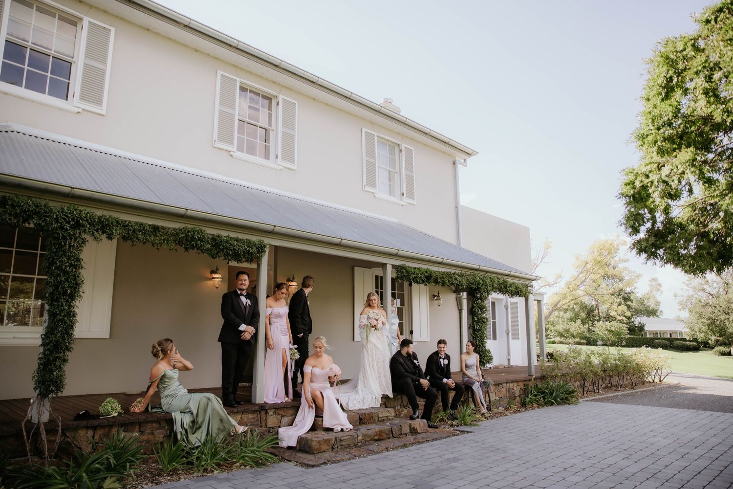 Wedding party gathered on the porch of a classic white colonial style house on a sunny afternoon.