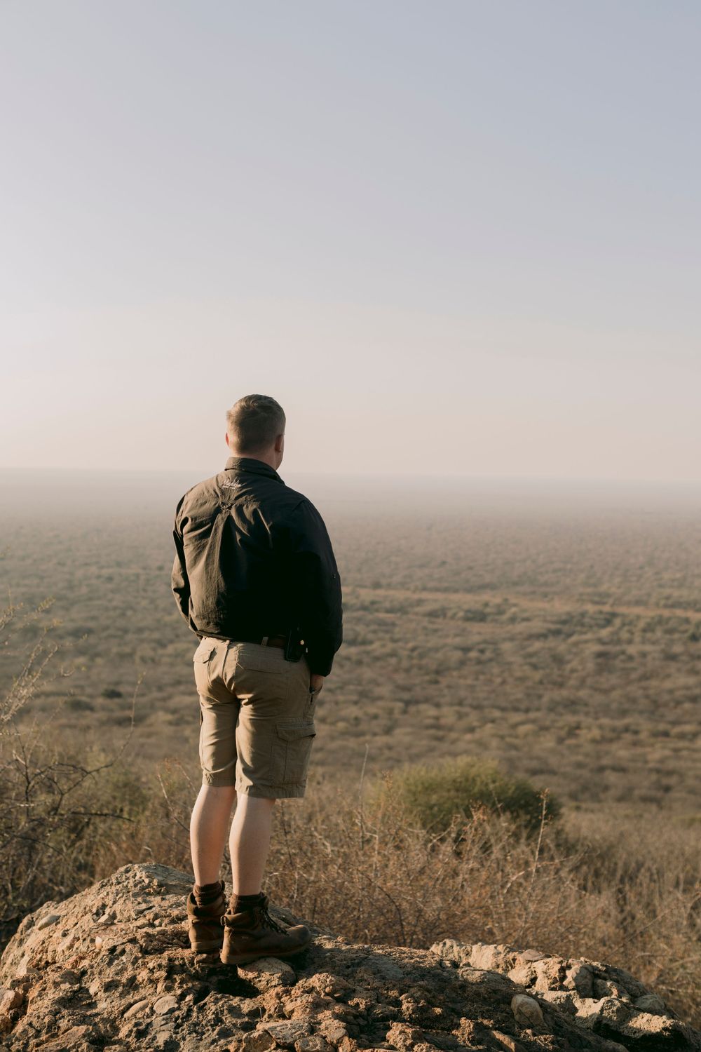 Man standing on rocky outcrop overlooking vast African savanna landscape at dusk.