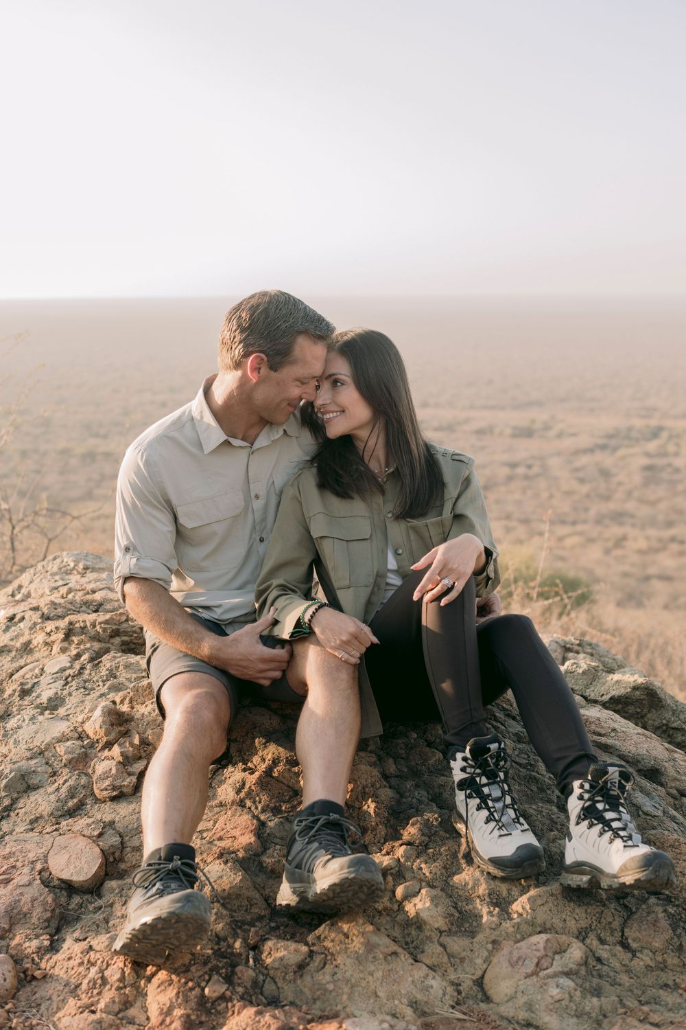Couple sitting closely on rocky hilltop with sweeping African savanna views behind them.