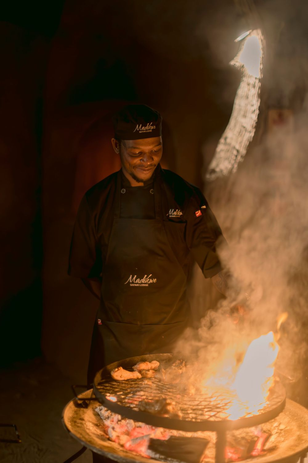 Chef grilling meat over open fire flames at a safari lodge outdoor kitchen.