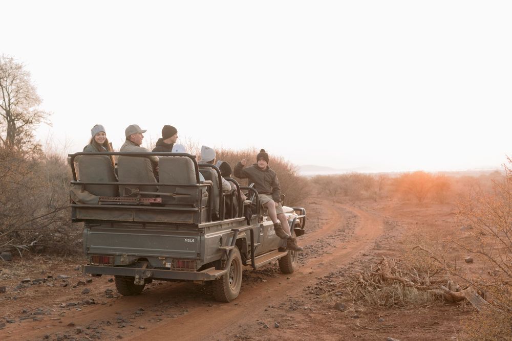 Group of tourists on a safari game drive vehicle traveling along a dusty dirt road at sunset.