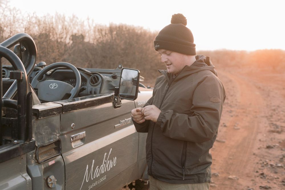 Man in winter hat and jacket checking his phone beside a safari game drive vehicle at sunset.