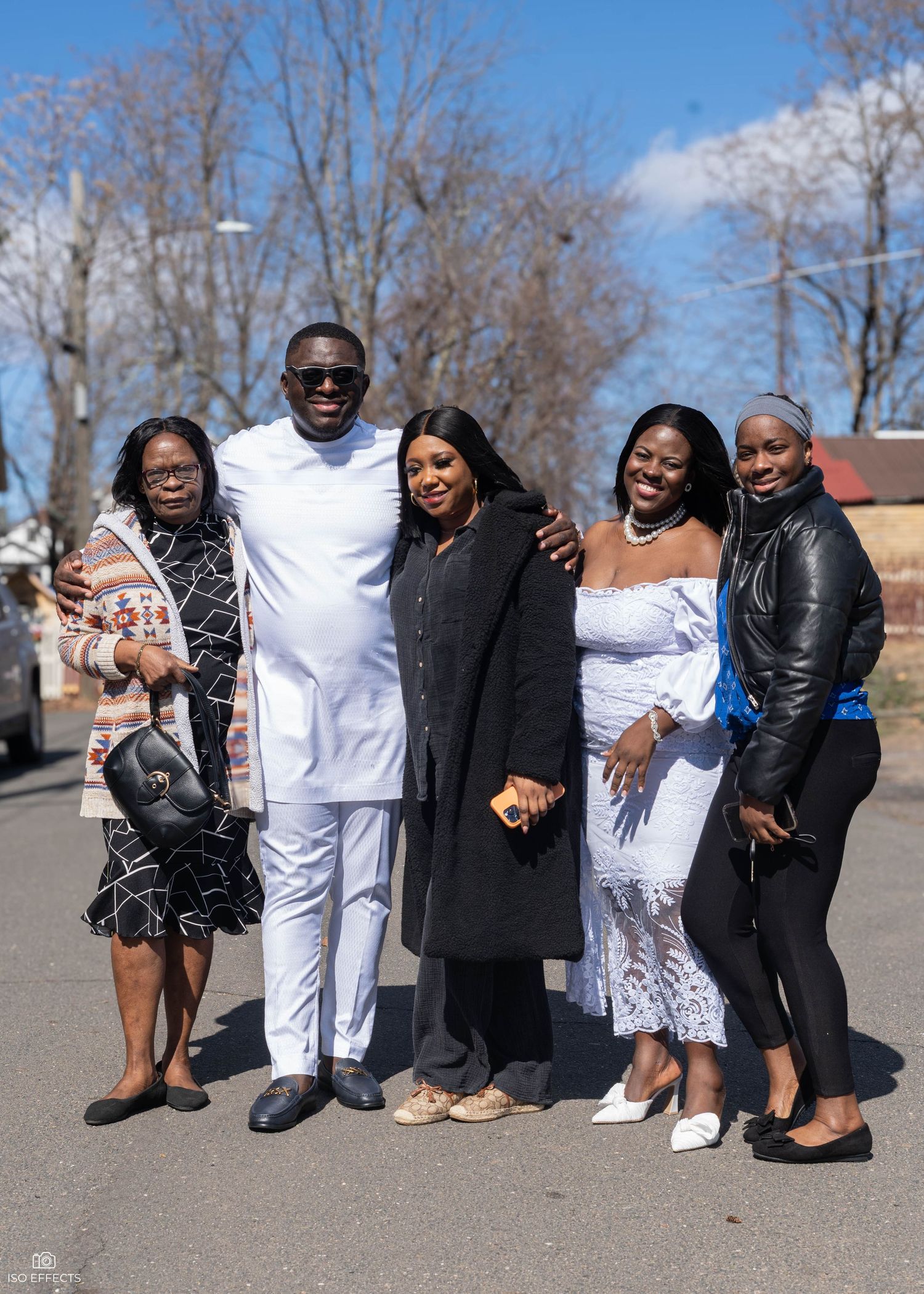 A group of people stands together outdoors on a sunny day, with one person wearing all white and others in dark attire.