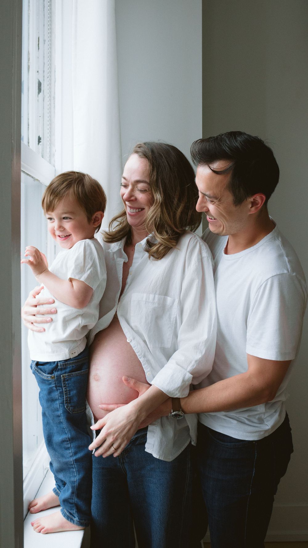 indoor family maternity photoshoot with the family laughing close to the window in matching outfits.