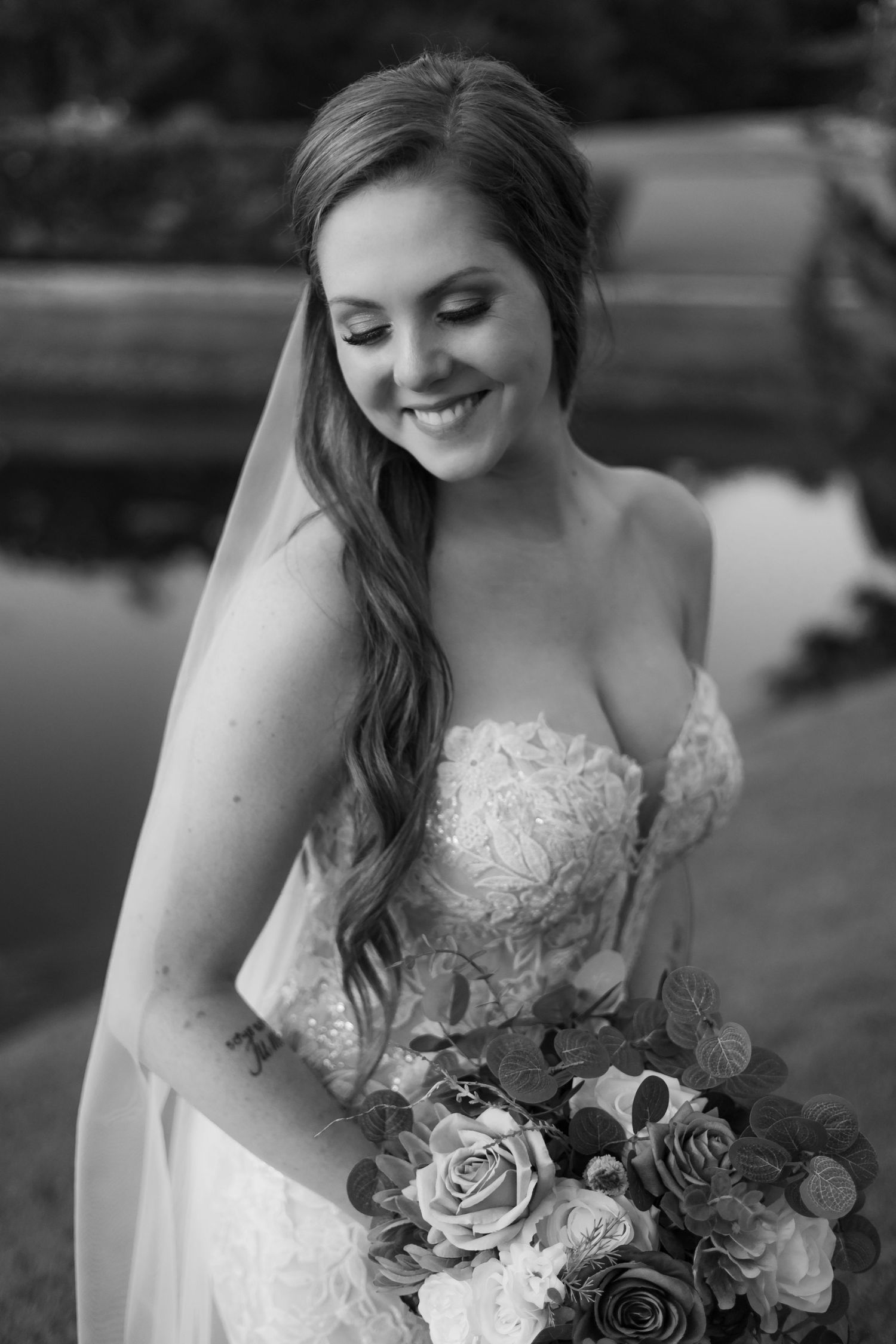 A bride in a lace wedding dress smiles while holding a beautiful bouquet of roses and mixed flowers.