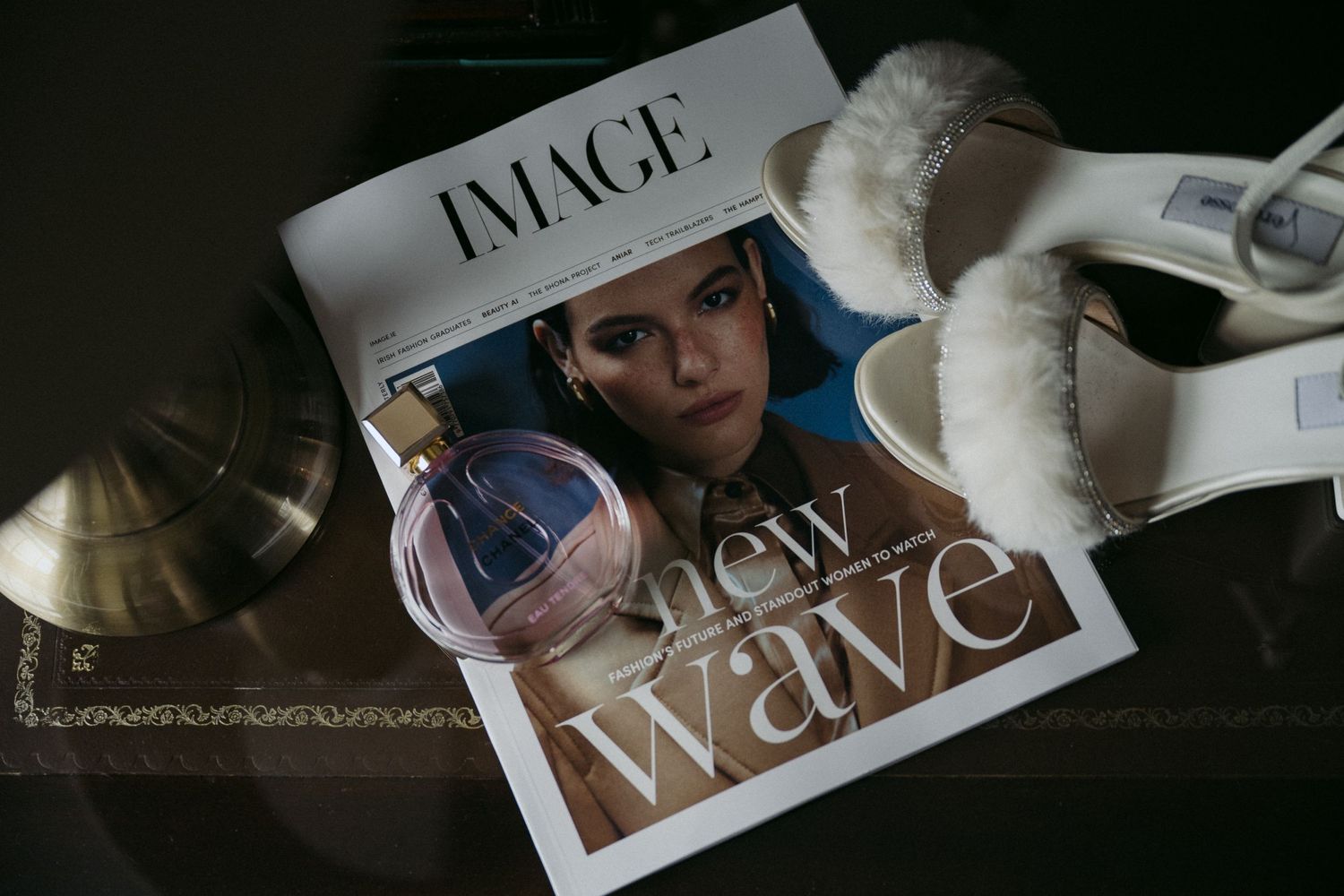 A lifestyle flatlay featuring a fashion magazine, perfume bottle, and white fluffy slippers on a dark surface.