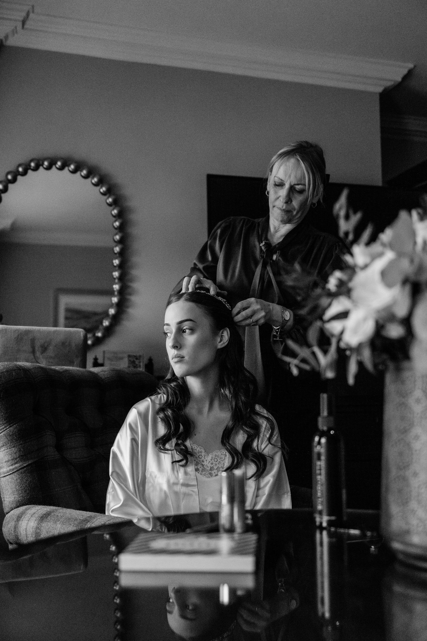A black and white photo showing preparation for a wedding as a stylist works on a bride's hair in an elegant room.