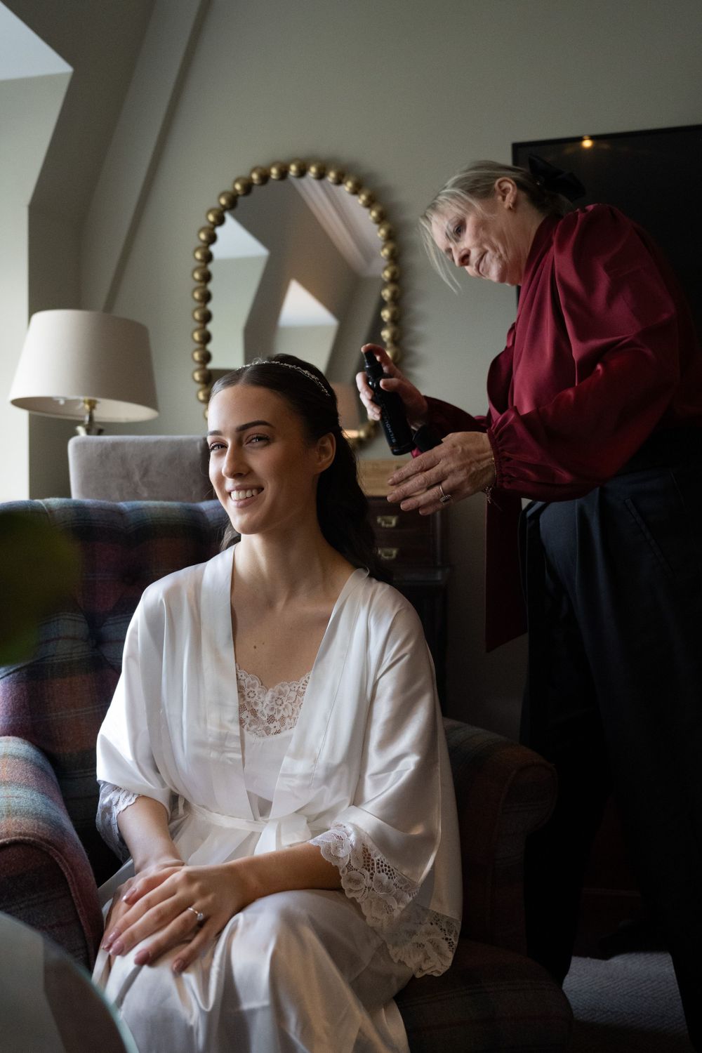 A stylist works on a client's hair as they sit in a white robe in a cozy room with a table lamp.