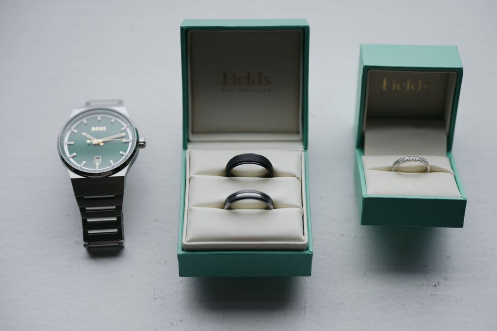 A stainless steel wristwatch displayed alongside two teal jewelry boxes containing rings on a light gray surface.
