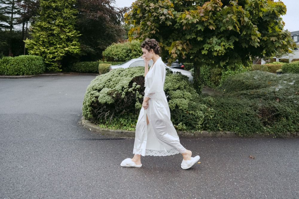 A person in a flowing white dress and sneakers walks gracefully on a winding path surrounded by lush green foliage.