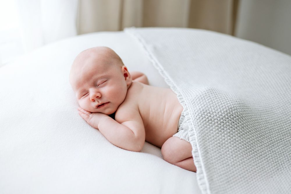 A peaceful sleeping baby lying his his front which his hand tucked under his chin and his bottom covered by a grey knit blanket.