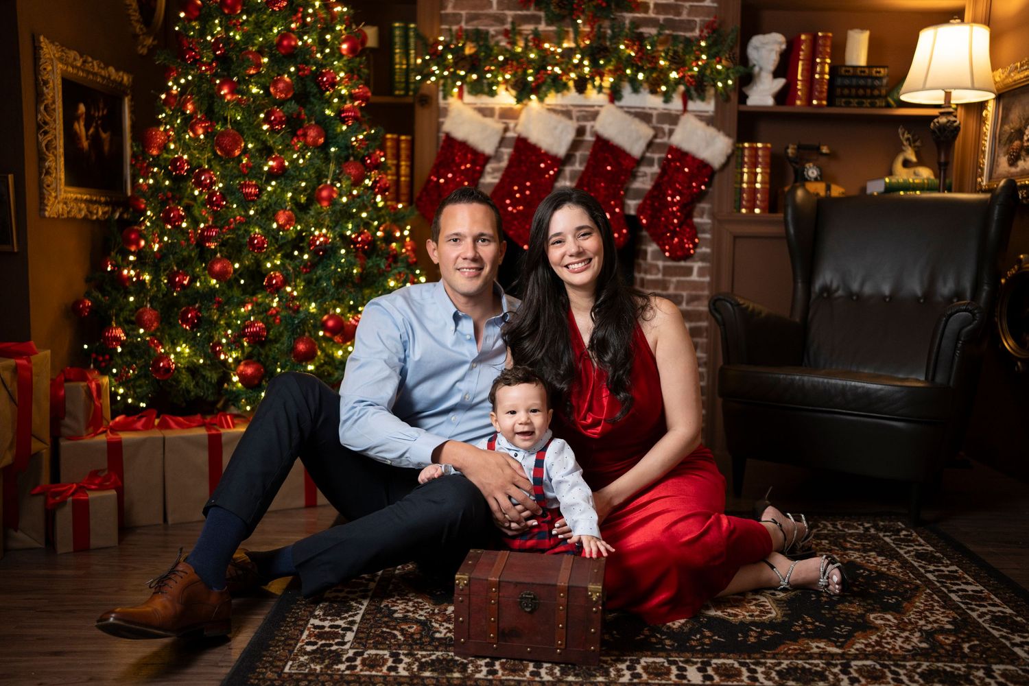 Family poses by Christmas tree with stockings and presents in cozy living room setting.