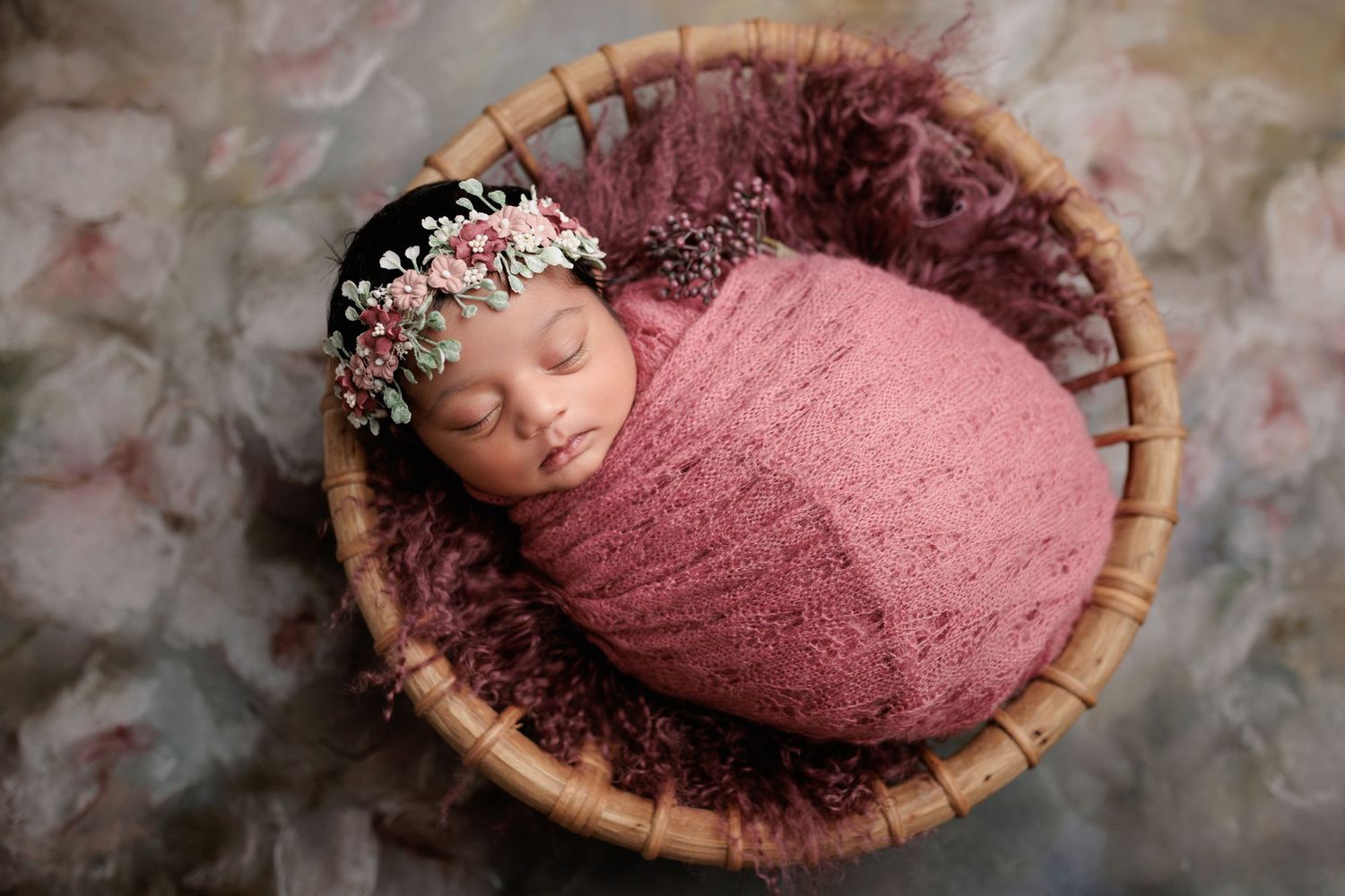 A sleeping newborn wrapped in a pink swaddle blanket rests in a round woven basket with a floral headpiece.