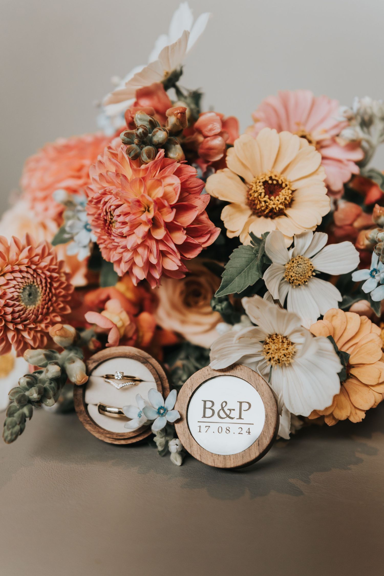 Wedding rings rest on wooden discs among coral dahlias and white zinnia flowers in romantic floral arrangement.