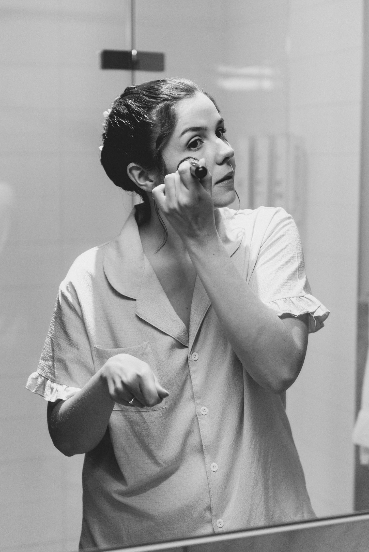 A person in a light colored button-down shirt poses expressively while applying makeup in a black and white photograph.