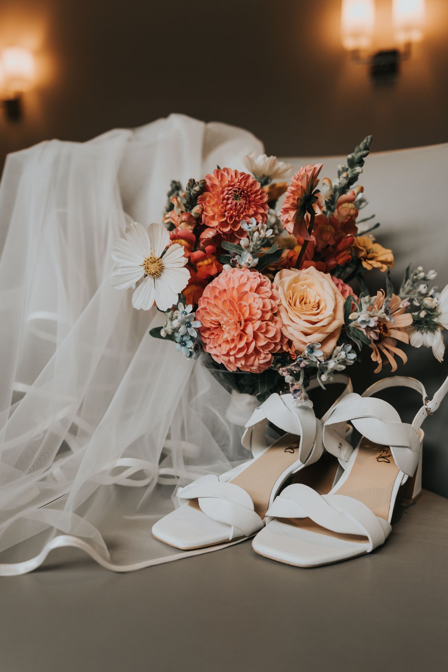 A bridal bouquet with coral carnations and white roses sits beside elegant white wedding shoes and flowing veil.