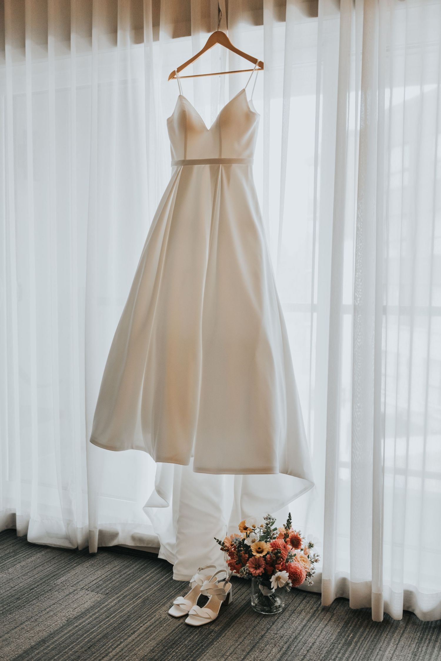 Elegant white wedding dress hangs by a window with a bridal bouquet of peach flowers and wedding shoes below.