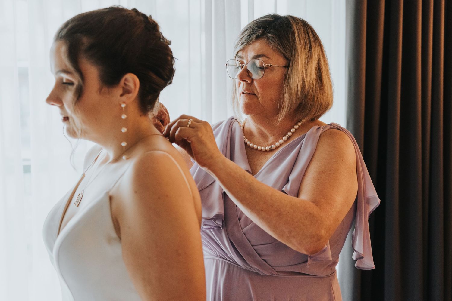 Someone helps button up a wedding dress in a soft natural light setting with white curtains in the background.